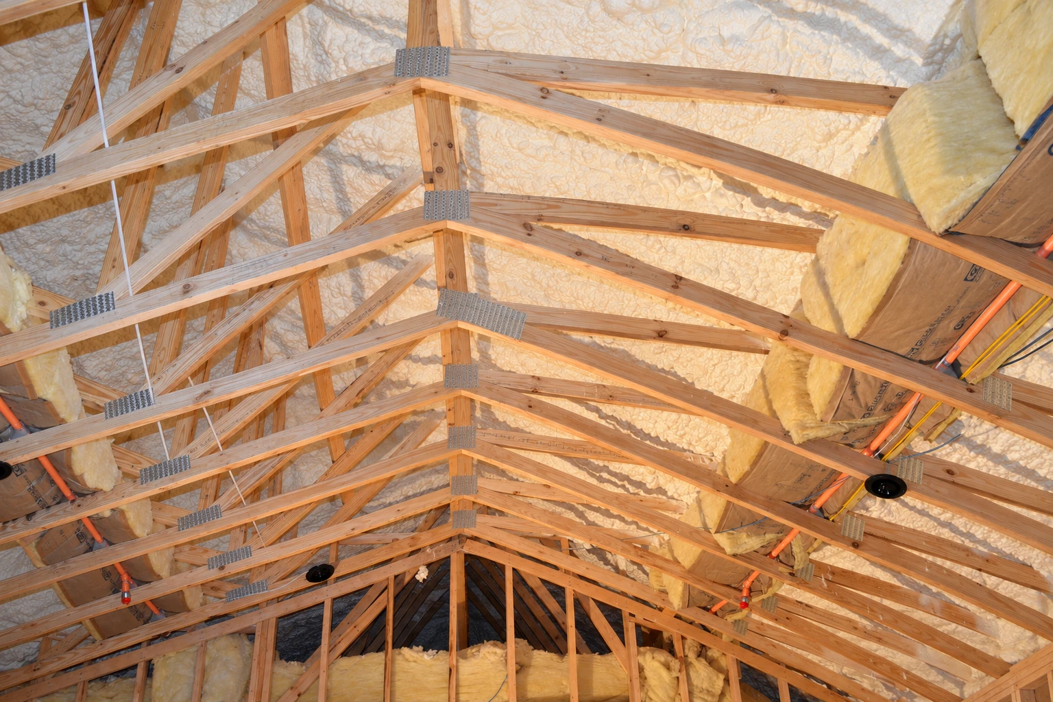 Wooden roof trusses frame an attic with spray foam on the roof and yellow batt insulation along the edges.