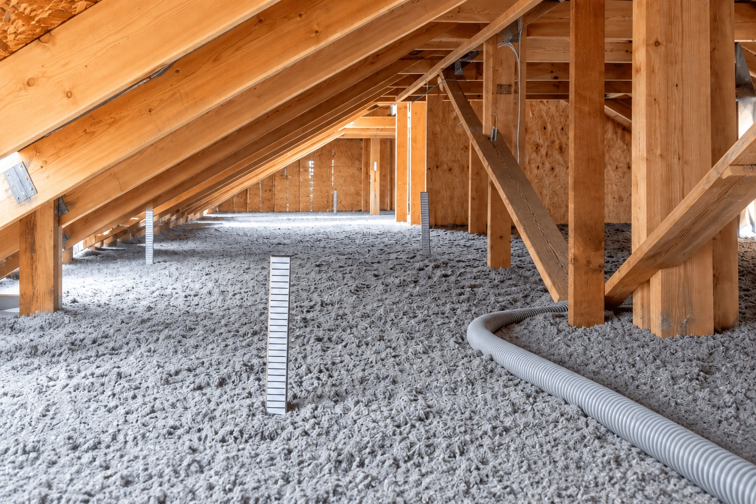 Attic floor covered in loose gray blown-in insulation, with a flexible hose extending across it.