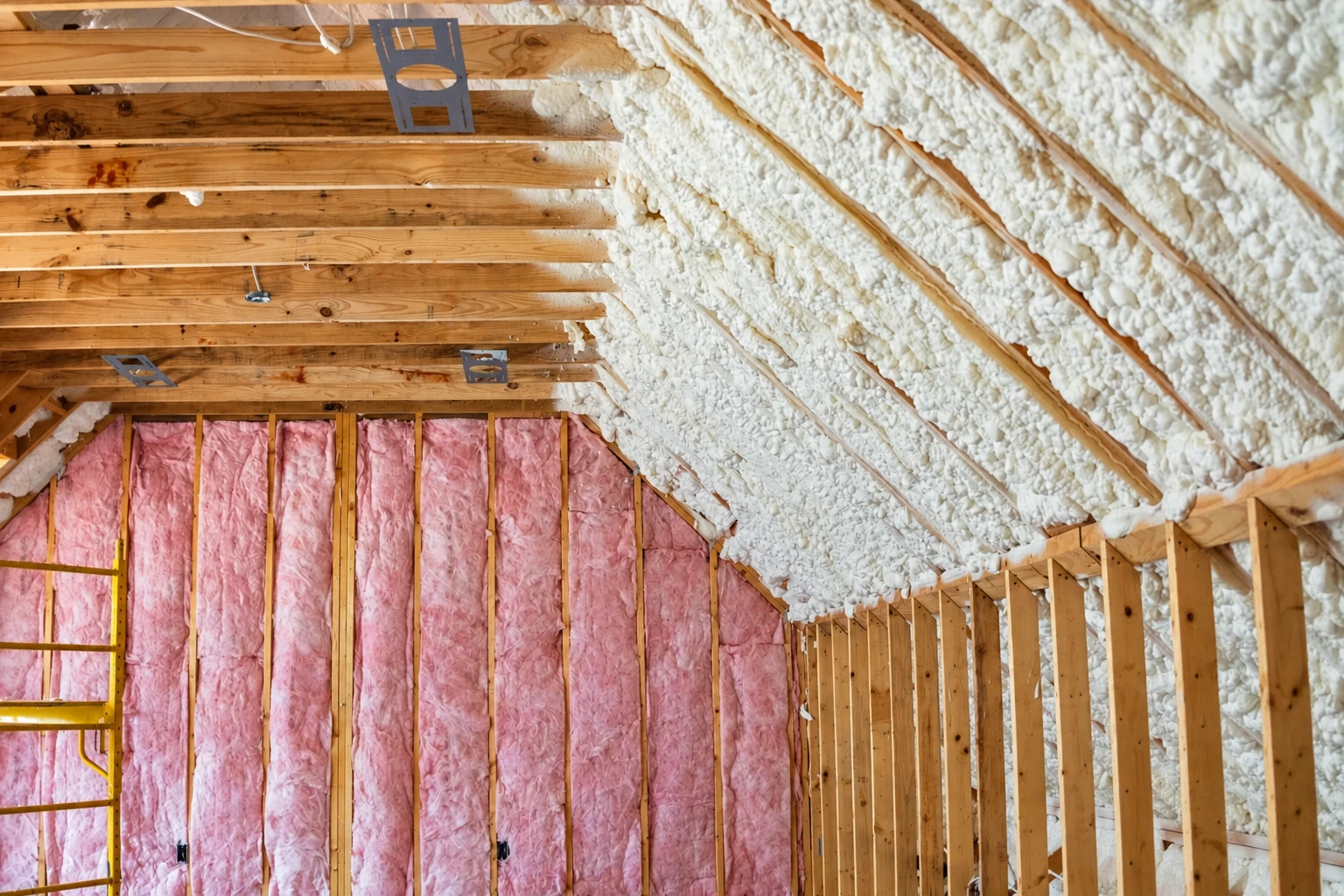 Open-cell foam coats the sloped roof between wooden rafters, above wall cavities filled with pink insulation.