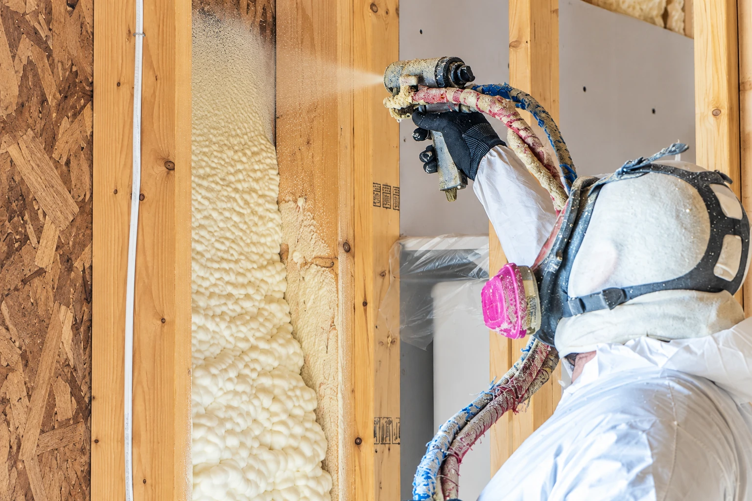 A worker in protective gear sprays foam insulation into an open wall cavity between wooden studs.
