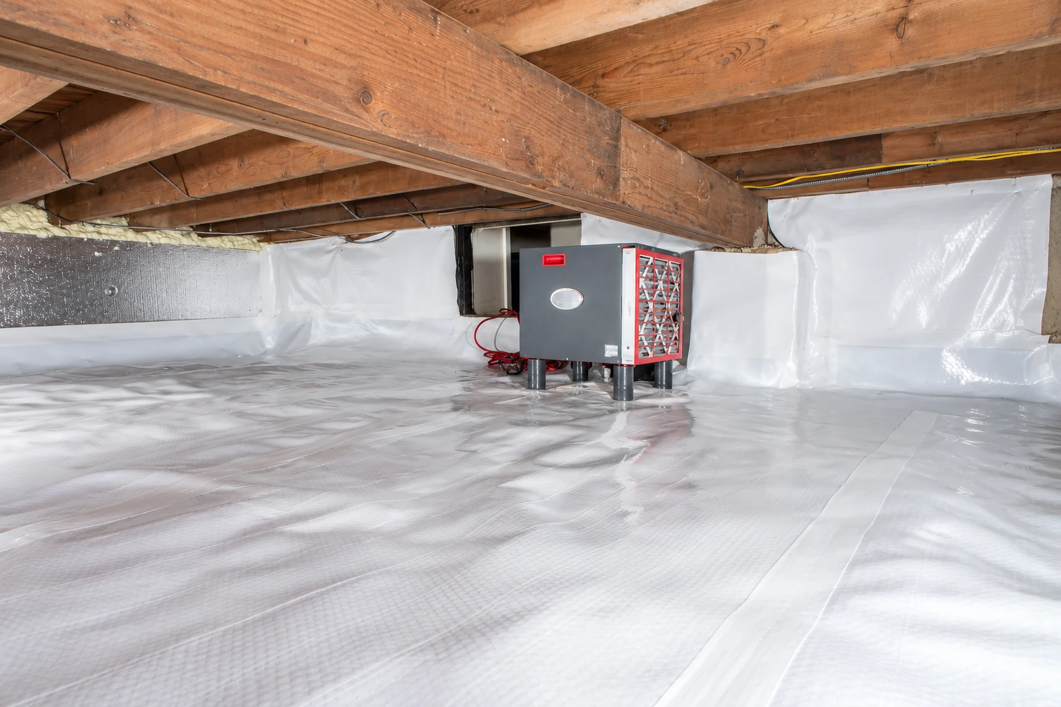 White vapor barrier covers a crawl space floor and lower walls around a red-and-gray mechanical unit beneath exposed wooden joists.