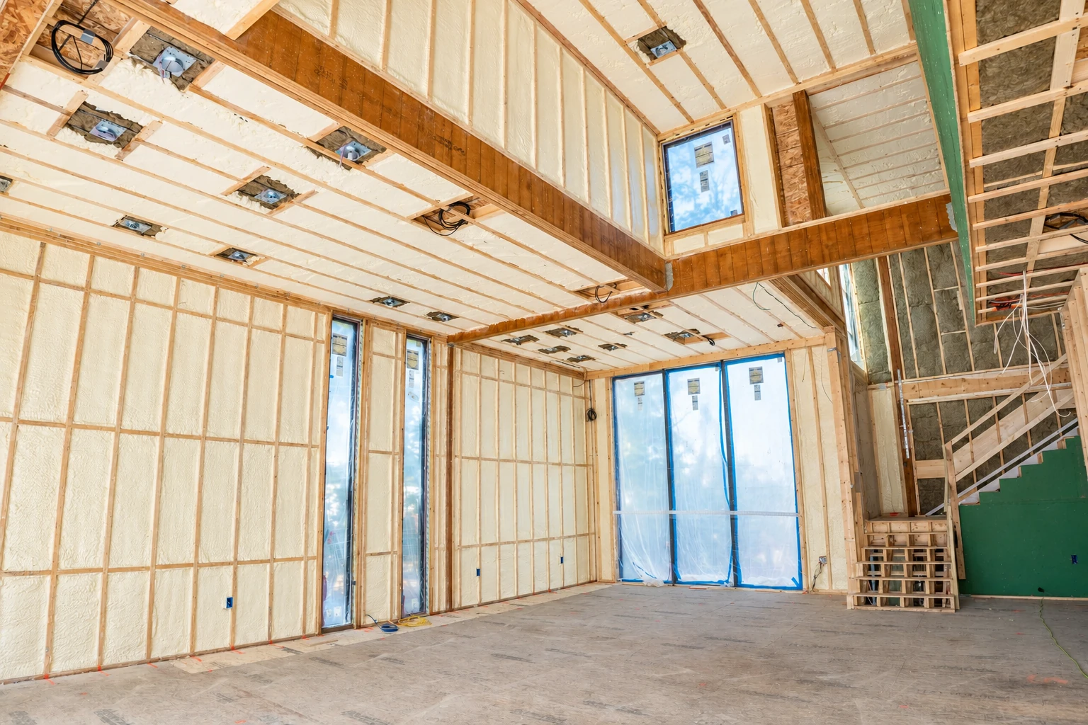 An unfinished house interior has pale spray foam insulation between wooden wall and ceiling framing.